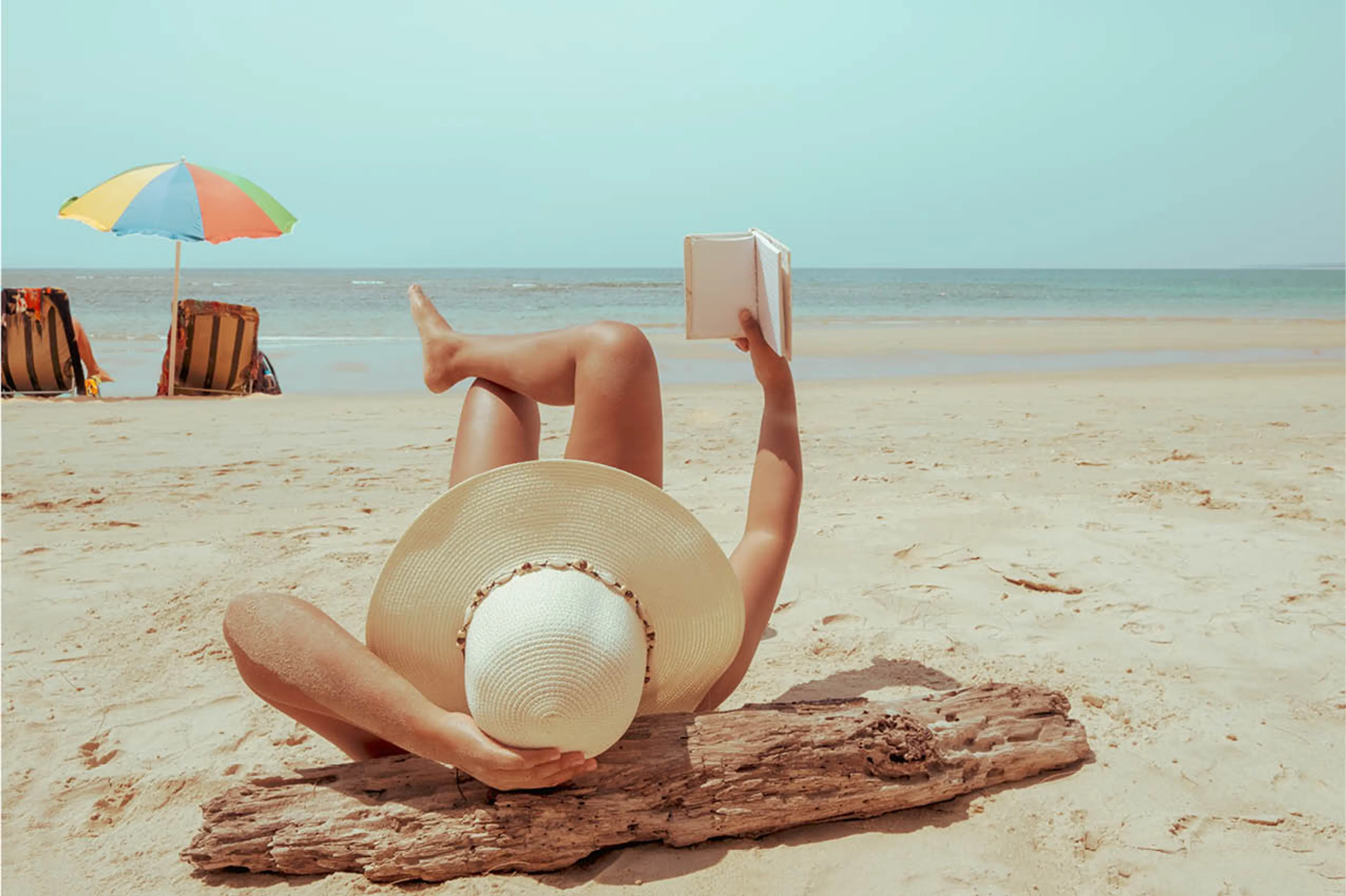 De vrouw op het strand ligt een boek te lezen. Er is niemand om haar heen en ze heeft een grote strandhoed op.